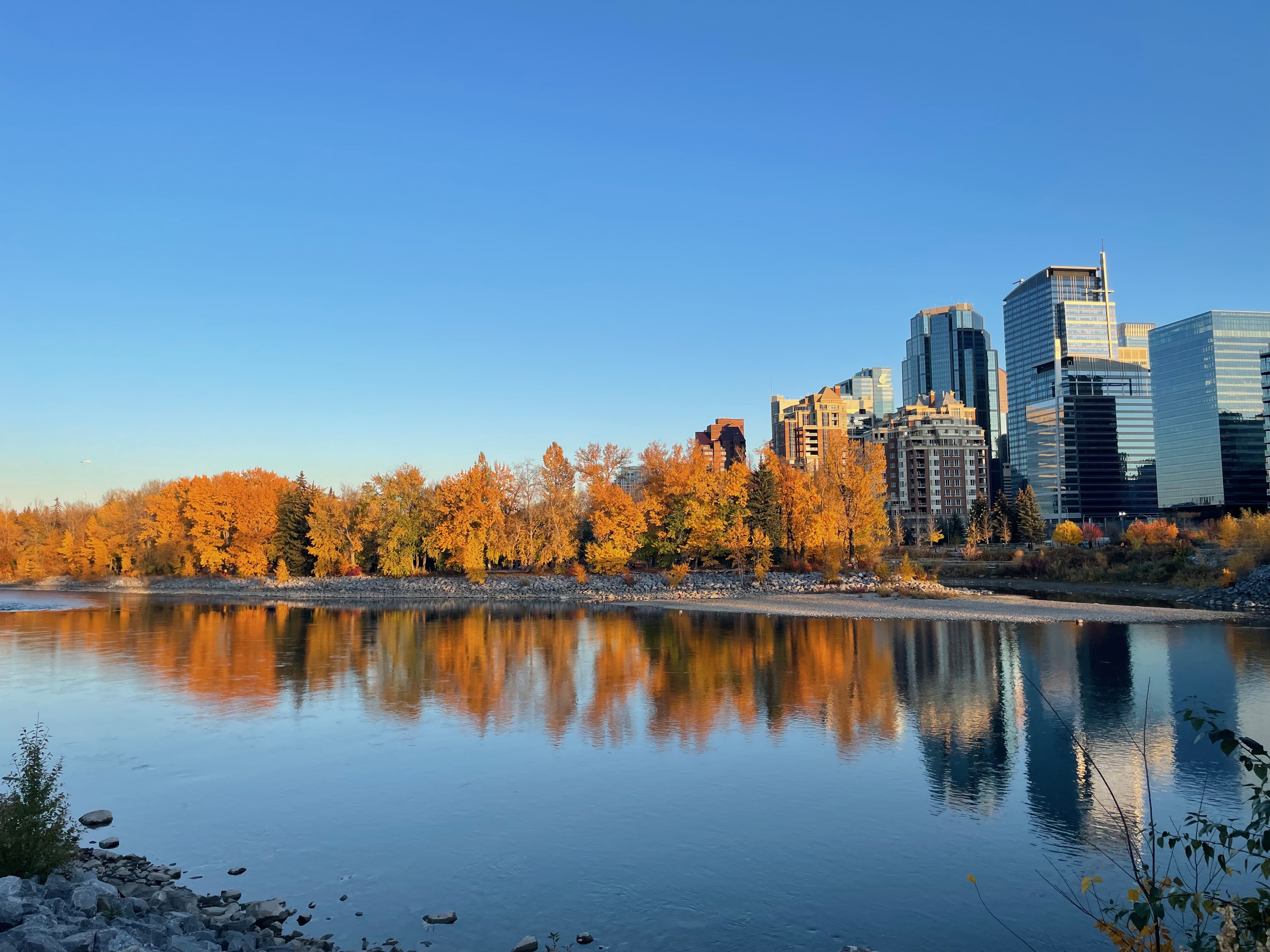 The image captures a scenic view of a river bordered by vibrant autumn foliage. On the left, clusters of trees display brilliant shades of orange and yellow, creating a warm, inviting atmosphere.