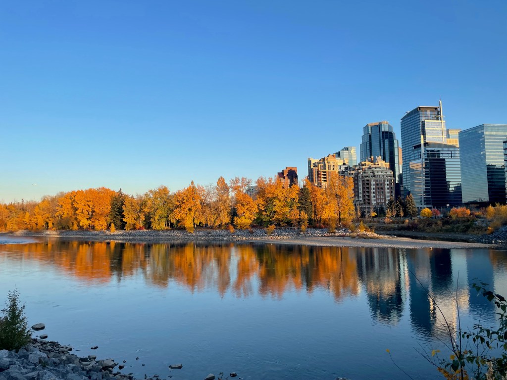 The image captures a scenic view of a river bordered by vibrant autumn foliage. On the left, clusters of trees display brilliant shades of orange and yellow, creating a warm, inviting atmosphere.