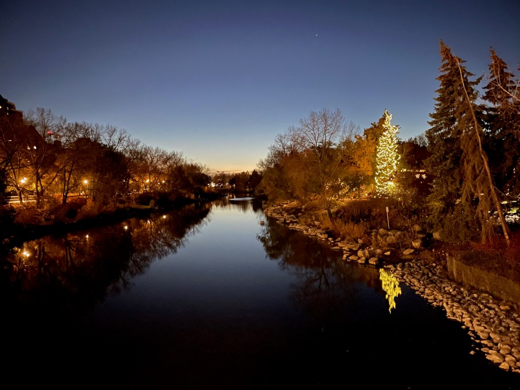 A calm river at dusk reflects dark trees, glowing park lights, and a brightly illuminated evergreen on the right bank. Bare branches and tall evergreens frame the water, while a deep blue evening sky with a few visible stars stretches overhead. Rocky shoreline and warm reflections create a peaceful winter scene.