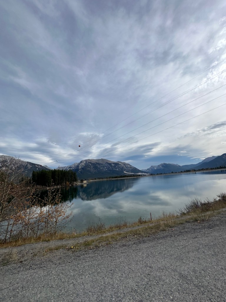 A calm mountain lake with trees, rocky peaks, and clouds reflected in the water.