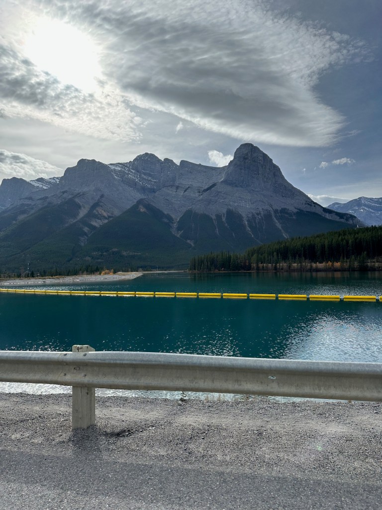 A turquoise mountain lake lies beneath towering gray peaks and dark evergreen forest. A line of bright yellow floating barriers crosses the water, while a metal roadside guardrail frames the foreground. Dramatic layered clouds stretch across the sky, casting soft shadows over the rugged landscape.