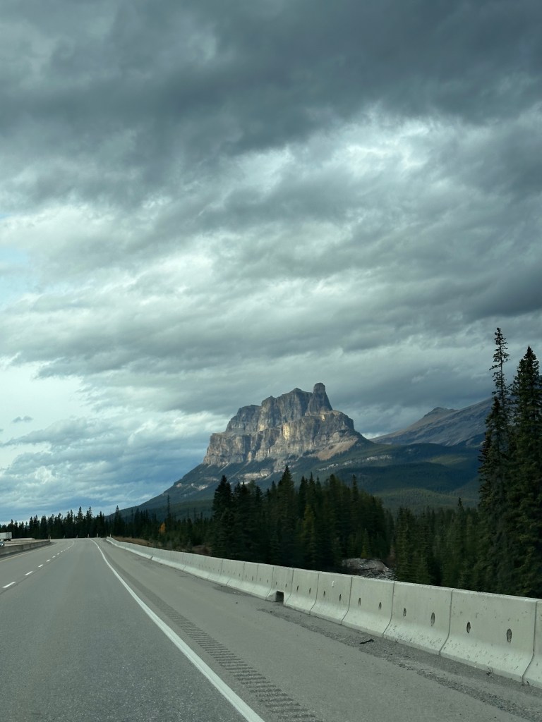 A highway through a forest leading toward a rocky mountain under storm clouds.