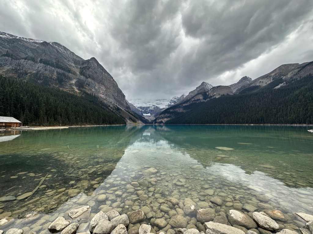 Clear lake water with stones, reflecting mountains, trees, and storm clouds.