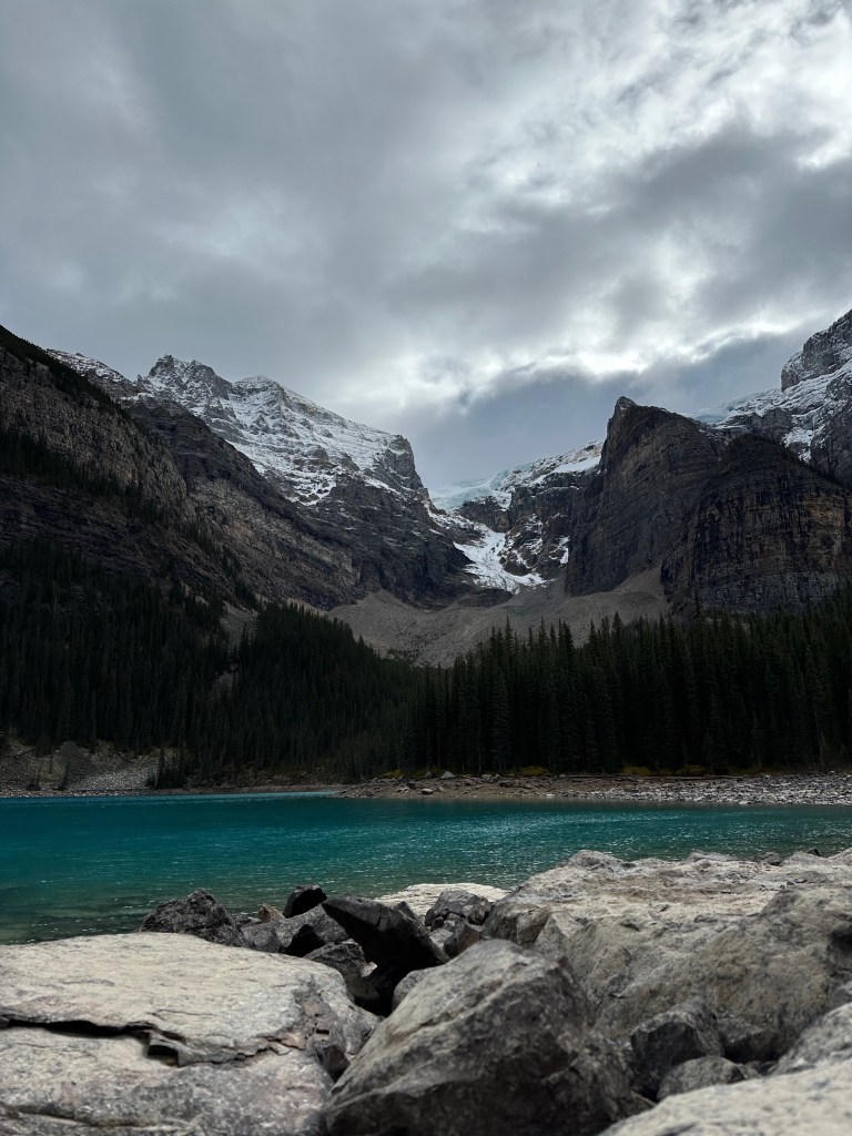 Large rocks along the shore of a turquoise mountain lake with evergreen trees and snow-covered peaks in the background.